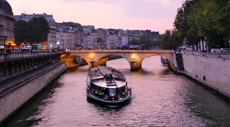 A Paris evening cruise boat passing under a glowing Seine bridge at twilight